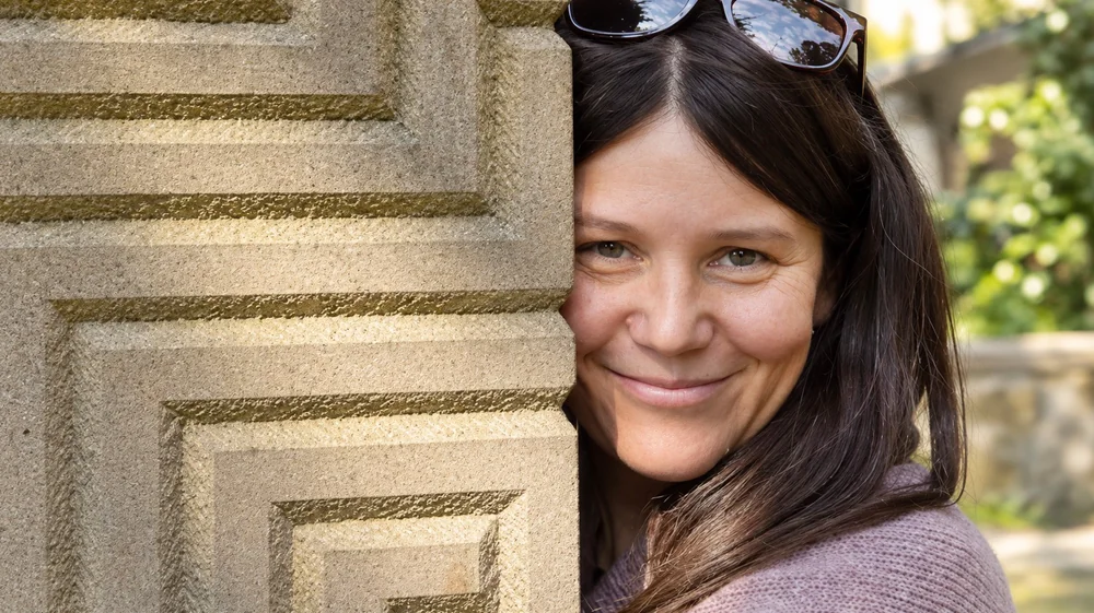 Daniela Kostinek blickt lächelnd hinter einer steinernen Säule hervor.
